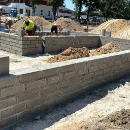 Motix - Wznoszenie ścian fundamentowych z bloczków betonowych na budowie domu jednorodzinnego. Widoczni pracownicy w trakcie murowania, hałdy piasku i materiały budowlane.