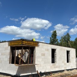 Motix - Wznoszenie ścian domu z bloczków Solbet, widoczne zbrojenie stropu i szalunki okienne. Na górze budynku pracownik w kamizelce odblaskowej. Błękitne niebo z chmurami w tle.