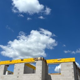 Motix - Wznoszenie ścian z betonu komórkowego na budowie domu jednorodzinnego, widok z dołu na fragment konstrukcji z żółtymi elementami szalunkowymi i błękitne niebo z chmurami.