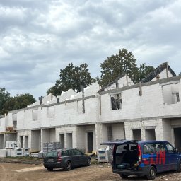 Motix - Wznoszenie szeregu domów z białego bloczka, widoczne zbrojenia i szalunki. W tle komin przemysłowy i pochmurne niebo. Na pierwszym planie zaparkowane samochody.