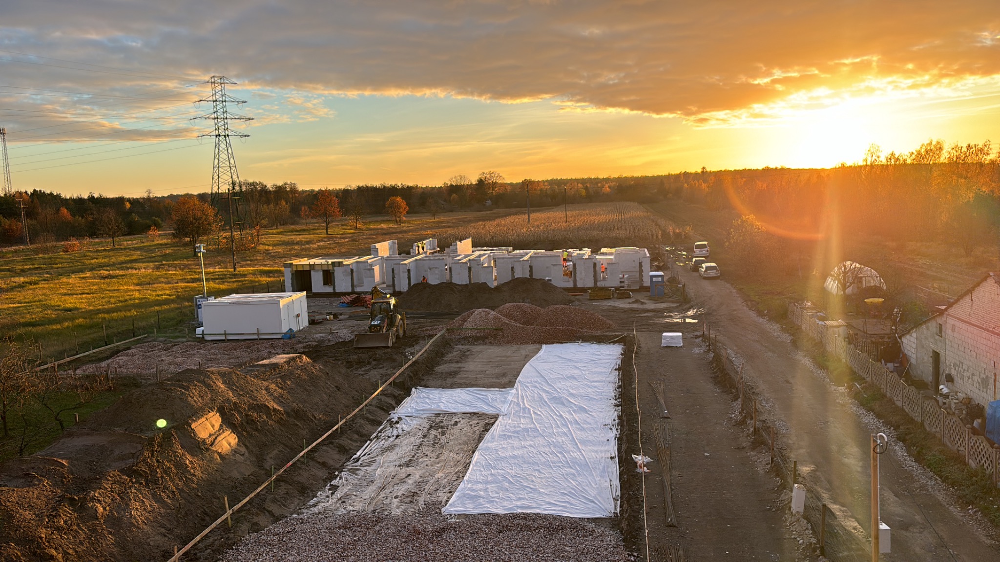 Budowa domu z betonu komórkowego w trakcie realizacji, widok z góry na fundamenty zabezpieczone folią, koparka i materiały budowlane na tle zachodzącego słońca.