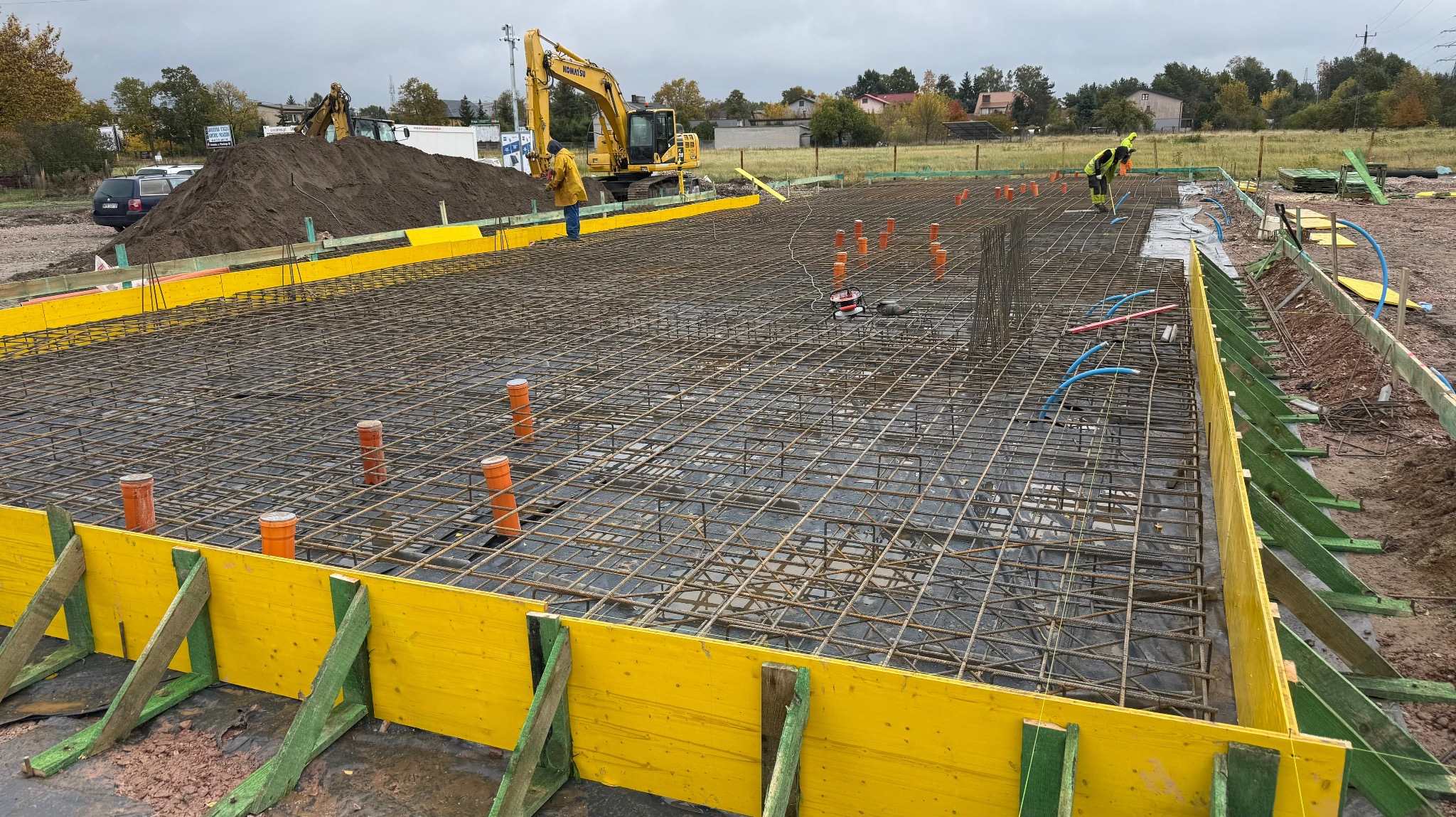 Zbrojenie fundamentu pod budynek z żółtym szalunkiem i wystającymi rurami. W tle koparka Komatsu i pracownicy budowlani. Widok na plac budowy w pochmurny dzień.