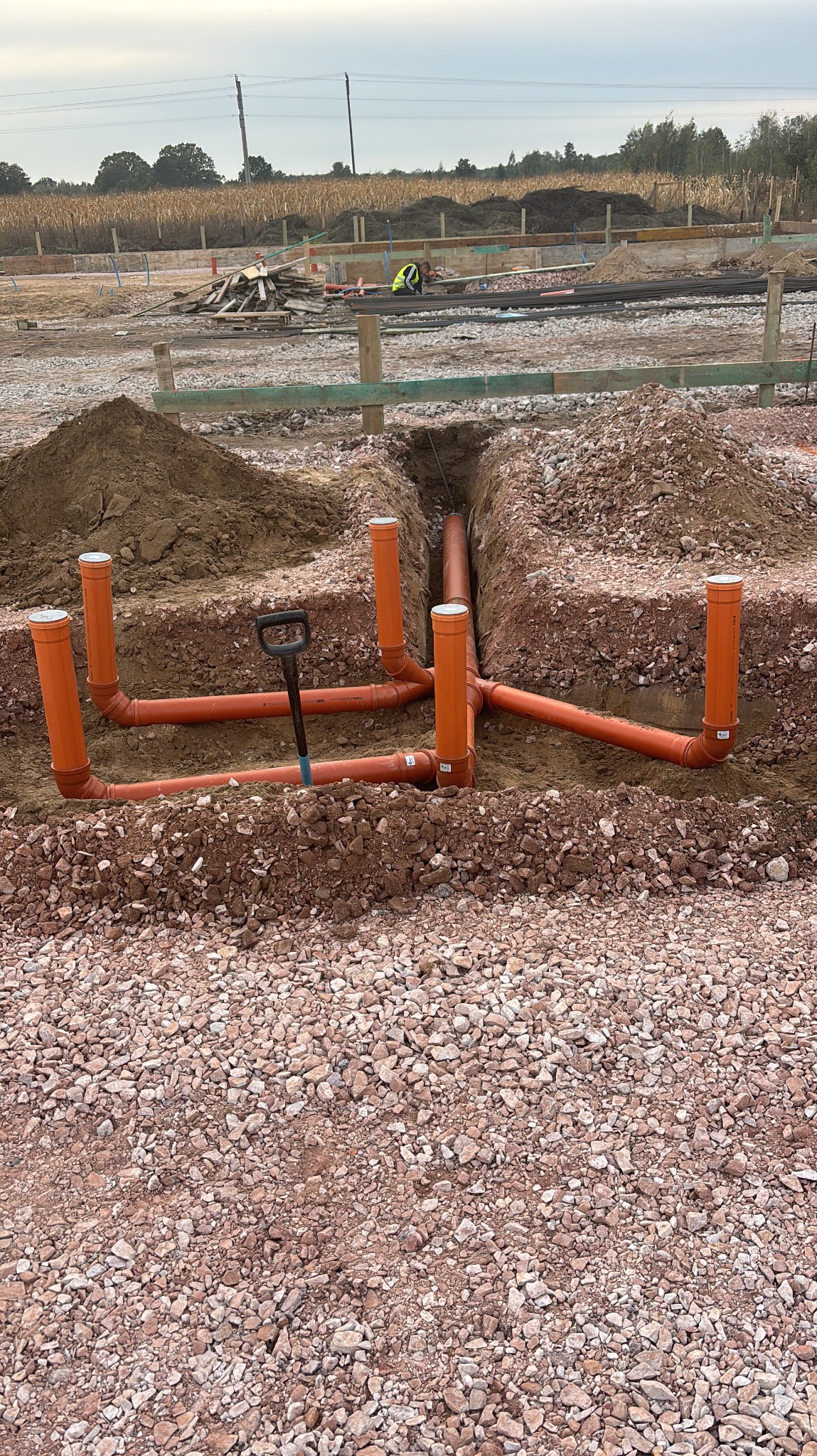 Pomarańczowe rury kanalizacyjne ułożone w wykopie, widoczna łopata. W tle teren budowy z drewnianymi elementami i pracującym robotnikiem w kamizelce.