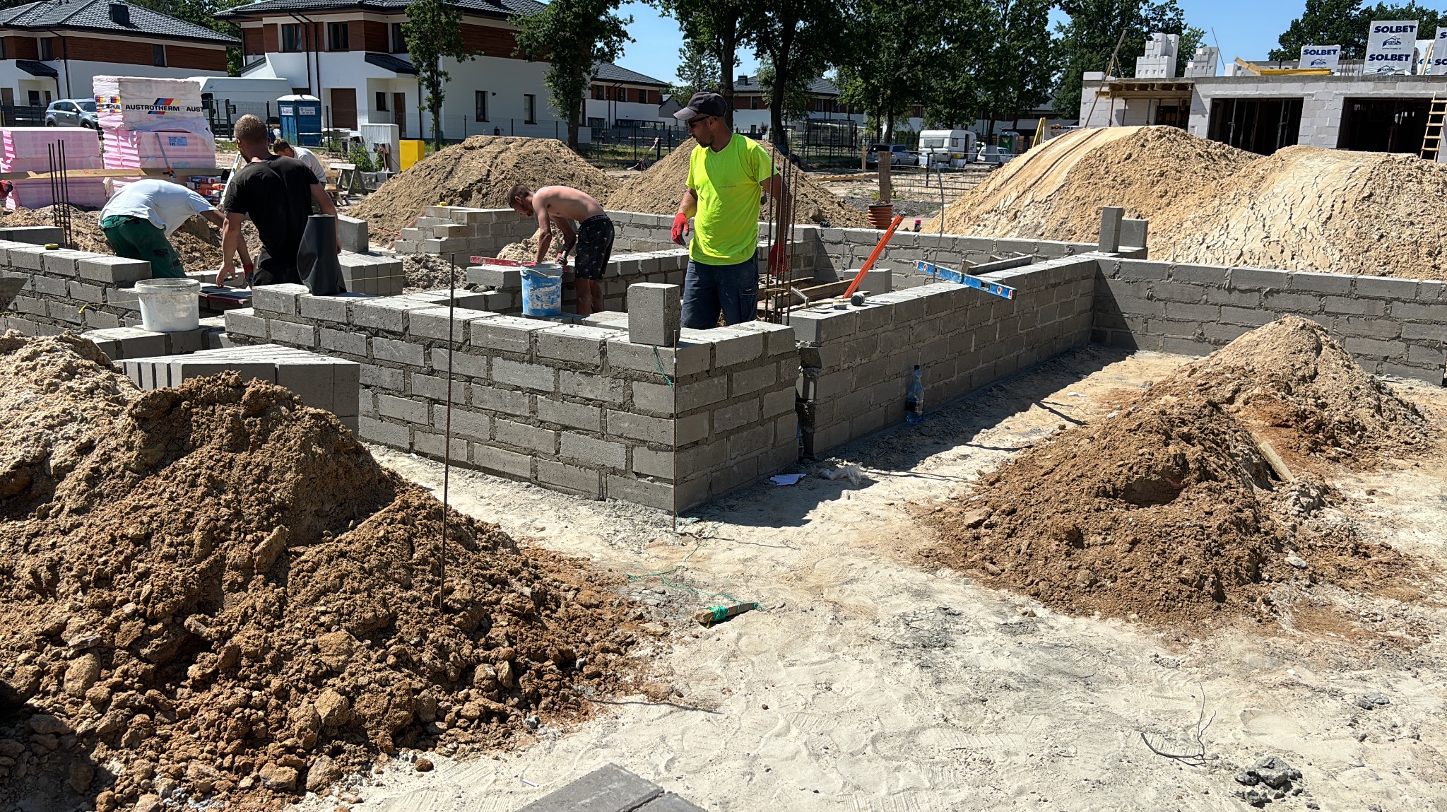 Wznoszenie fundamentów z bloczków betonowych. Murarze w trakcie pracy, widoczne narzędzia i materiały budowlane. Na drugim planie domy w zabudowie jednorodzinnej.