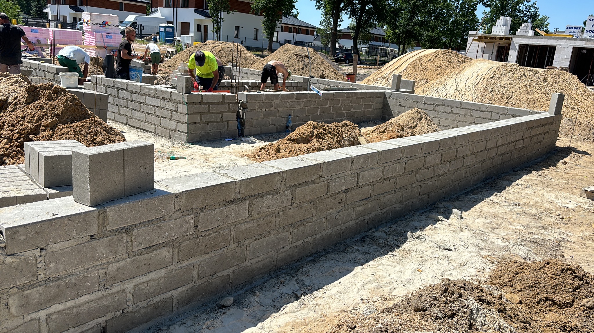 Wznoszenie ścian fundamentowych z bloczków betonowych na budowie domu jednorodzinnego. Widoczni pracownicy w trakcie murowania, hałdy piasku i materiały budowlane.