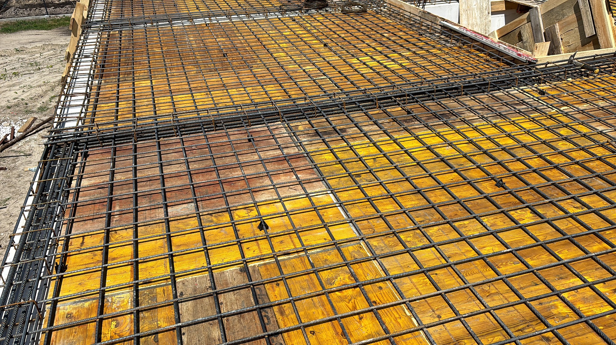 Zbrojenie płyty fundamentowej na deskowaniu z żółtej sklejki. Widok z góry, siatka z prętów stalowych ułożona krzyżowo. Elementy drewniane szalunku widoczne w tle.