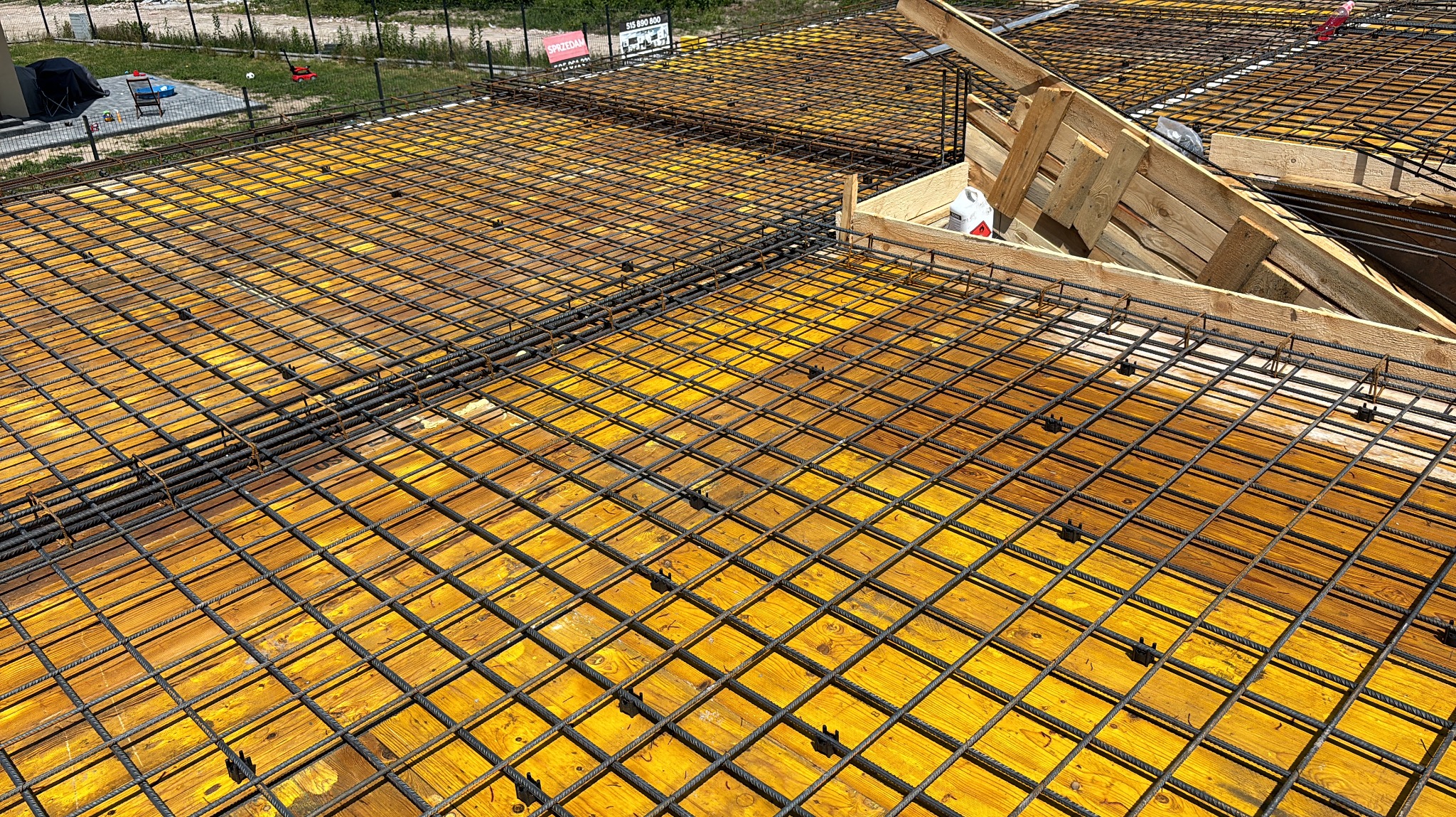 Zbrojenie płyty fundamentowej z żółtym szalunkiem, widok z góry. Stalowe pręty tworzą regularną siatkę. W tle widoczne elementy drewniane i zieleń.