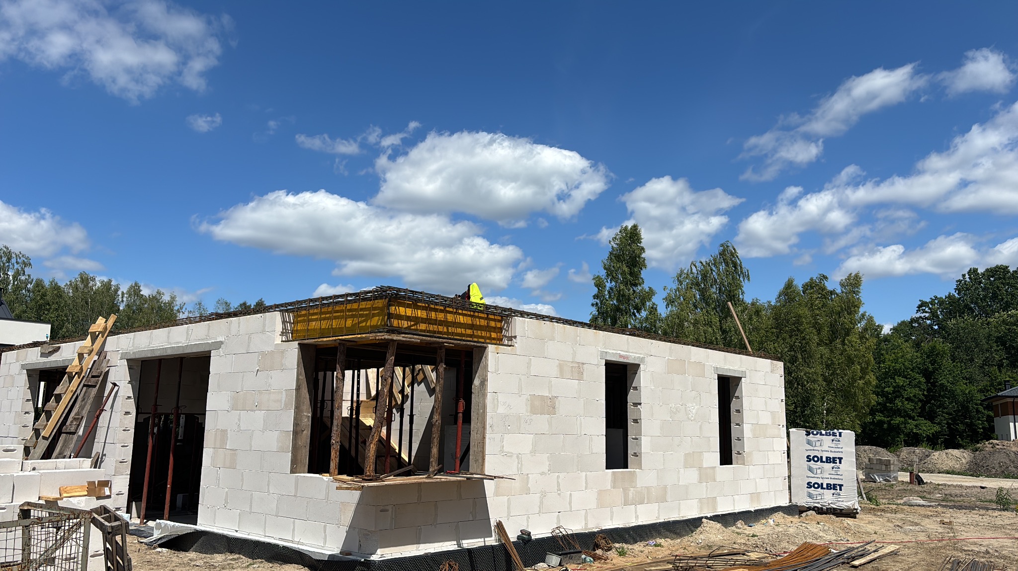 Wznoszenie ścian domu z bloczków Solbet, widoczne zbrojenie stropu i szalunki okienne. Na górze budynku pracownik w kamizelce odblaskowej. Błękitne niebo z chmurami w tle.