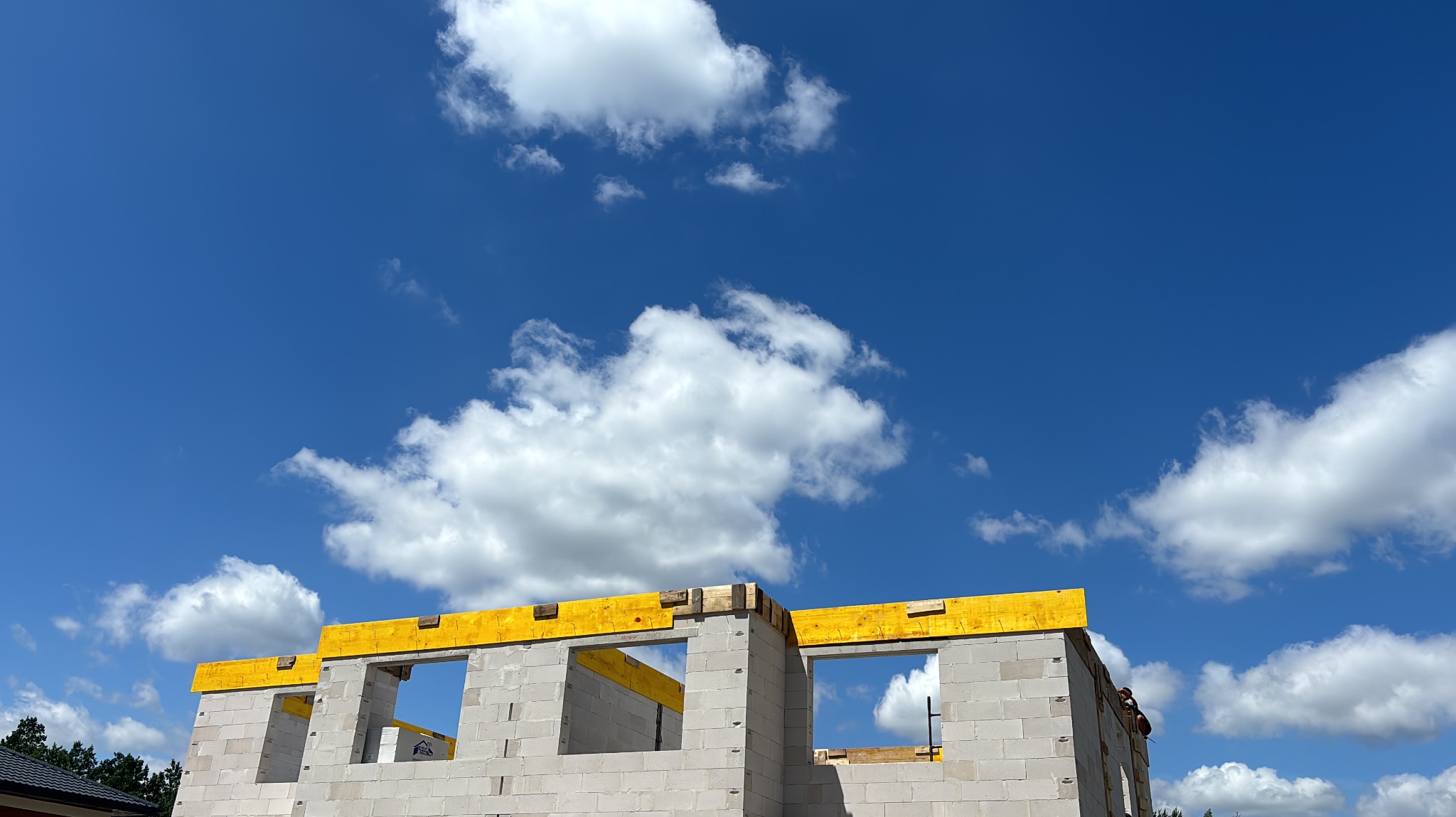 Wznoszenie ścian z betonu komórkowego na budowie domu jednorodzinnego, widok z dołu na fragment konstrukcji z żółtymi elementami szalunkowymi i błękitne niebo z chmurami.