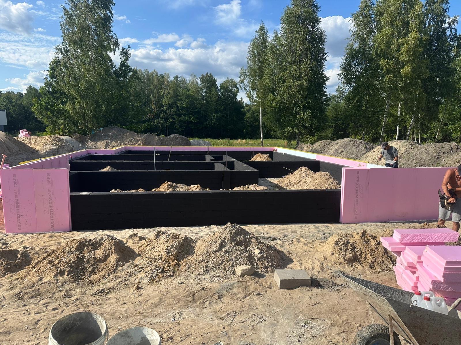 Fundamenty domu jednorodzinnego z izolacją termiczną z różowych płyt Austrotherm XPS, widoczne zbrojenia i piasek wewnątrz. Prace budowlane w toku, słoneczny dzień.