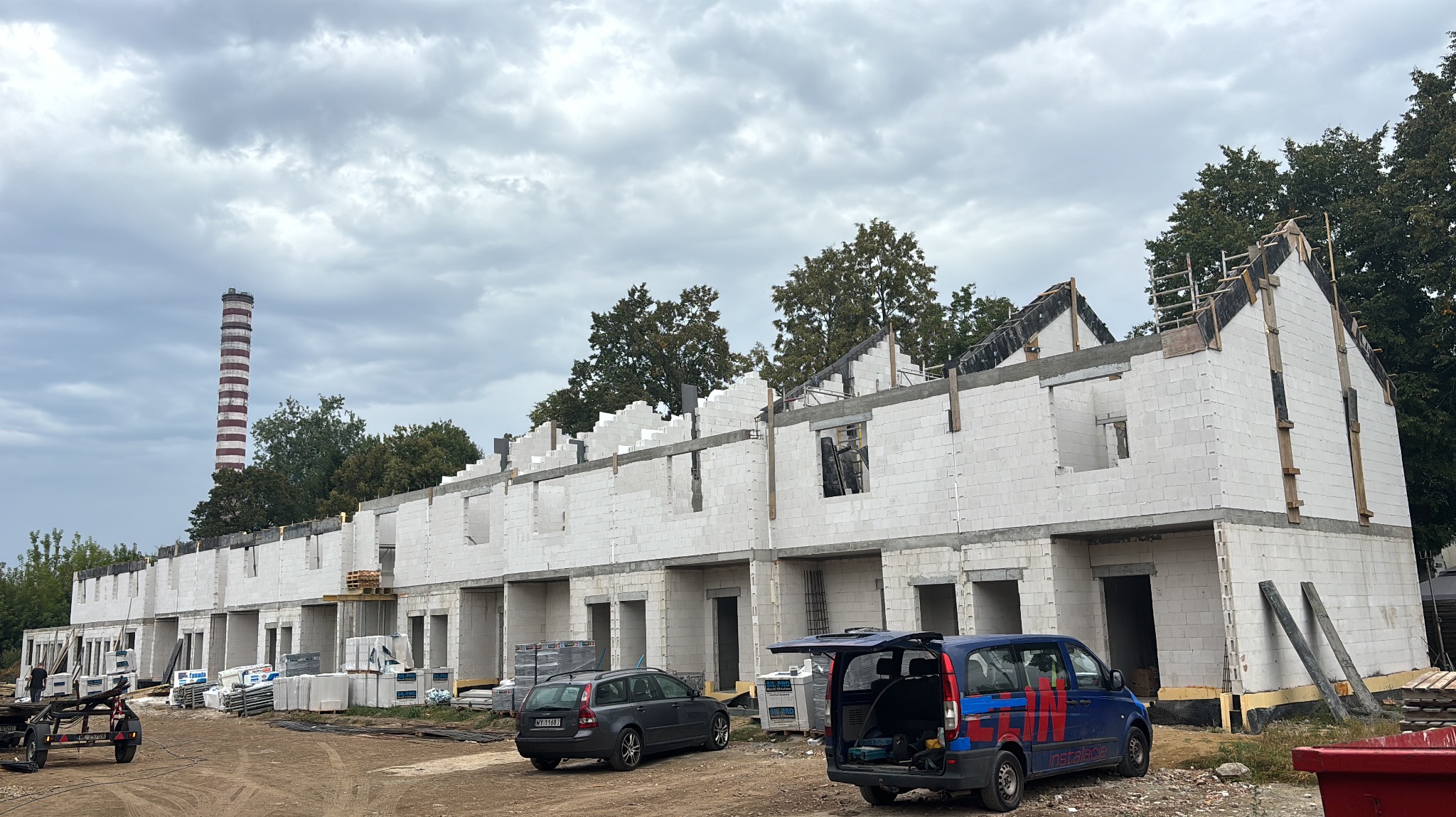 Wznoszenie szeregu domów z białego bloczka, widoczne zbrojenia i szalunki. W tle komin przemysłowy i pochmurne niebo. Na pierwszym planie zaparkowane samochody.