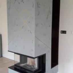 Nowoczesny kominek narożny z szarą obudową zdobioną złotymi geometrycznymi liniami, czarnym wkładem i przeszklonym paleniskiem, usytuowany w jasnym wnętrzu.