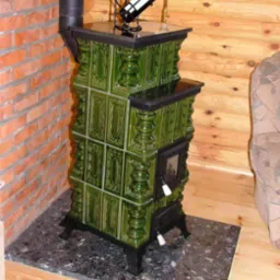 Zielony, kaflowy piec z czarnym blatem i rurą dymową, stojący na marmurowej podstawie w rogu drewnianego domu, z dekoracyjną butelką wina na górze.