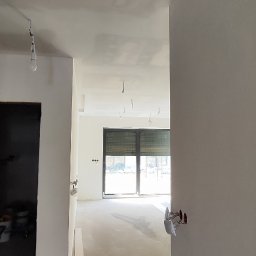 remontEX KRZYSZTOF CHYLA - Wnętrze w trakcie wykończenia: gładkie, białe ściany, surowa podłoga, widoczne punkty elektryczne. Widok na okno balkonowe z żaluzjami. Częściowo widoczny sufit z punktami do oświetlenia.