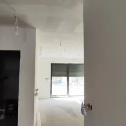 Wnętrze w trakcie wykończenia: gładkie, białe ściany, surowa podłoga, widoczne punkty elektryczne. Widok na okno balkonowe z żaluzjami. Częściowo widoczny sufit z punktami do oświetlenia.