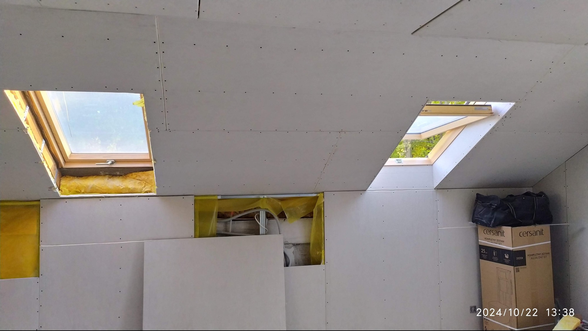 Montaż płyt karton-gips na poddaszu z oknami dachowymi, widoczne izolacje i instalacje. Częściowo zabudowane ściany, karton Cersanit w rogu.
