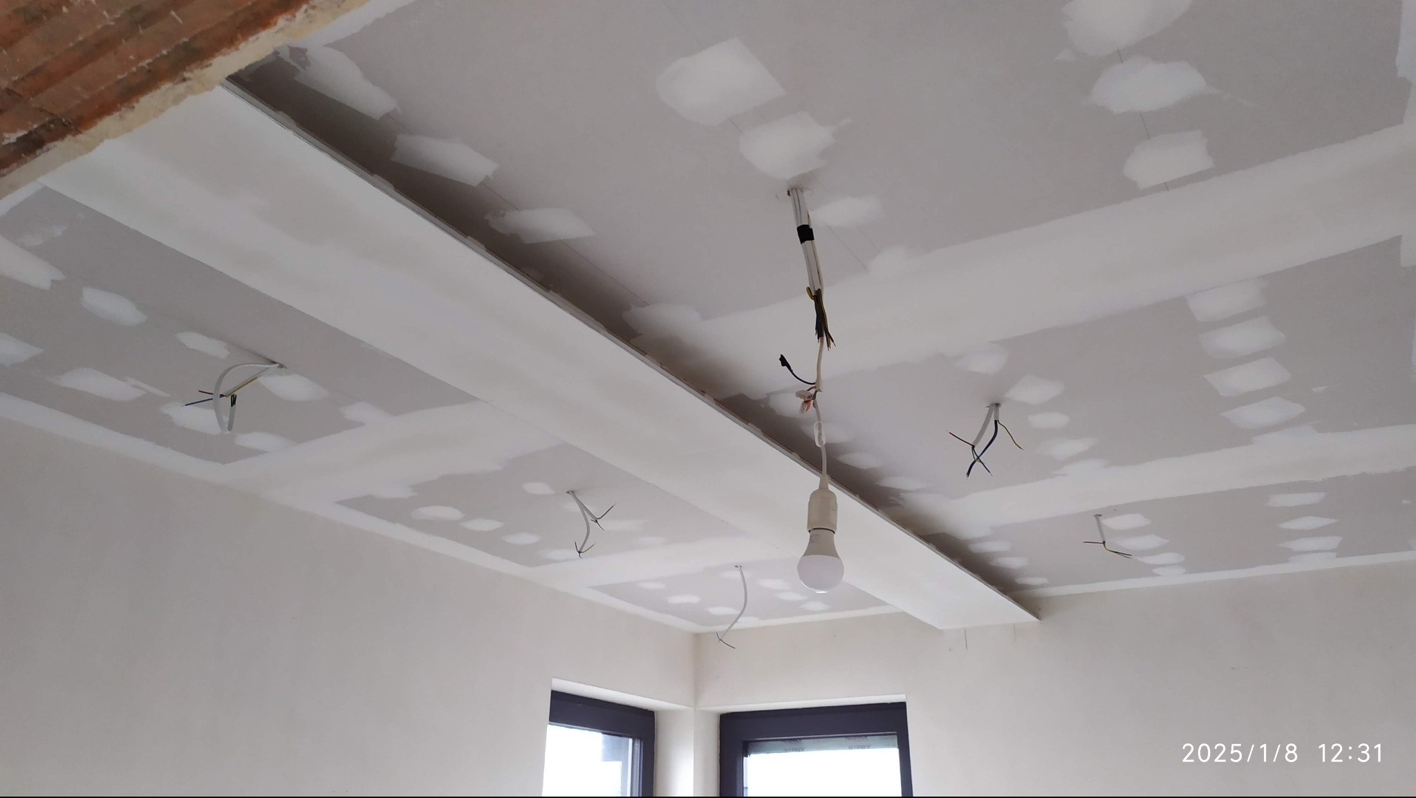 Sufit z płyt kartonowo-gipsowych w trakcie montażu, widoczne kable oświetleniowe. Na suficie widoczne miejsca szpachlowania łączeń płyt. W tle okno.