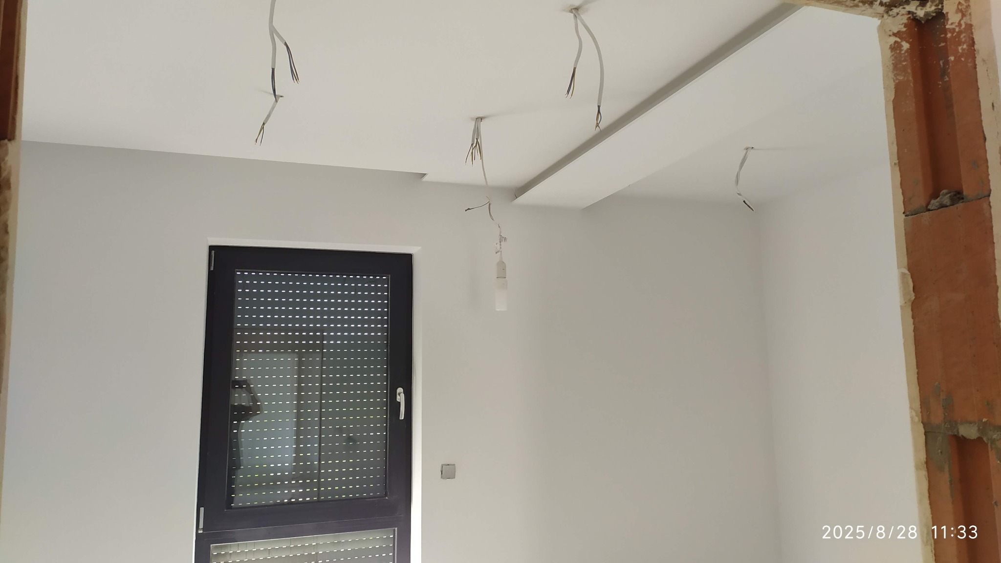 Białe, świeżo pomalowane wnętrze z częściowo zamontowaną płytą karton-gips na suficie. Widoczne wystające kable elektryczne i fragment ościeżnicy z cegły.