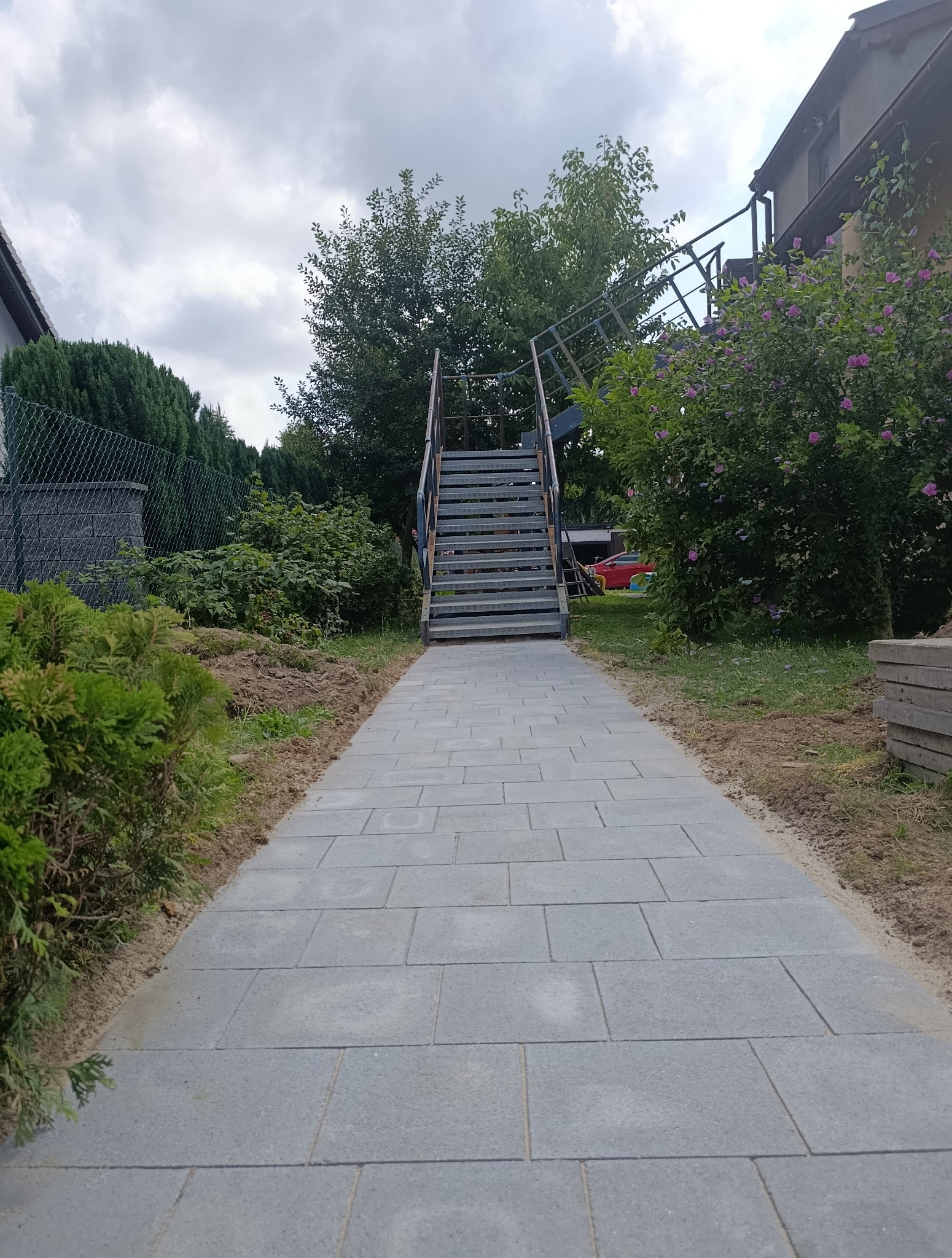 Ułożona ścieżka z szarej kostki brukowej prowadząca do schodów na zewnątrz budynku, otoczona zielenią i fragmentem ogrodzenia.