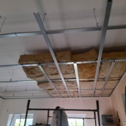 Fachouse - Montaż izolacji z wełny mineralnej w metalowym stelażu sufitu podwieszanego. Widoczne profile i fragment rusztowania.