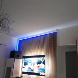 Nowoczesny salon z drewnianą ścianą lamelową podświetloną niebieskim LED, telewizor, kryształowy żyrandol i białe meble w minimalistycznym stylu.