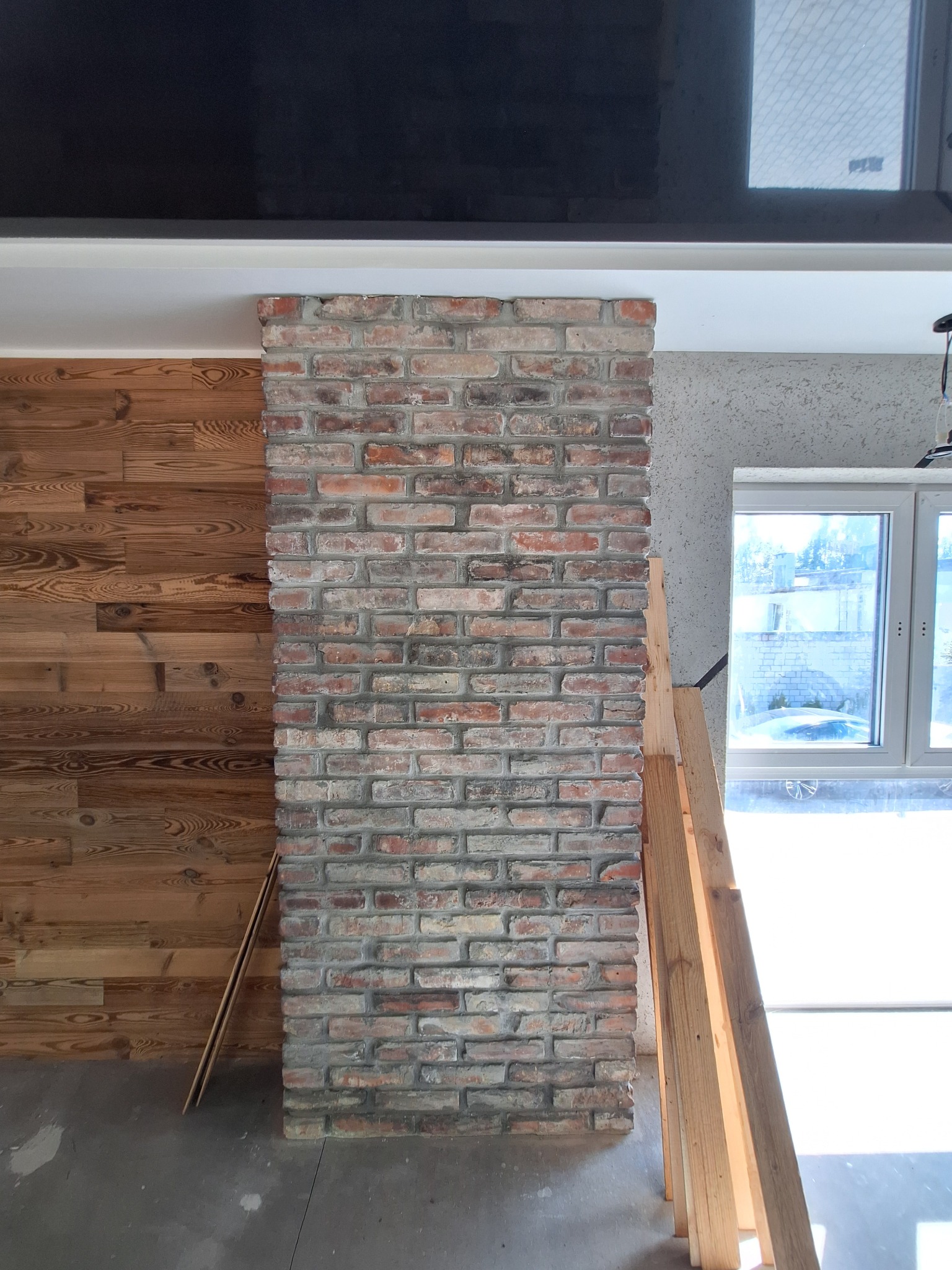 Wnętrze z ceglaną ścianą i drewnianą boazerią, w trakcie prac wykończeniowych. Widoczne deski i surowe podłoże.