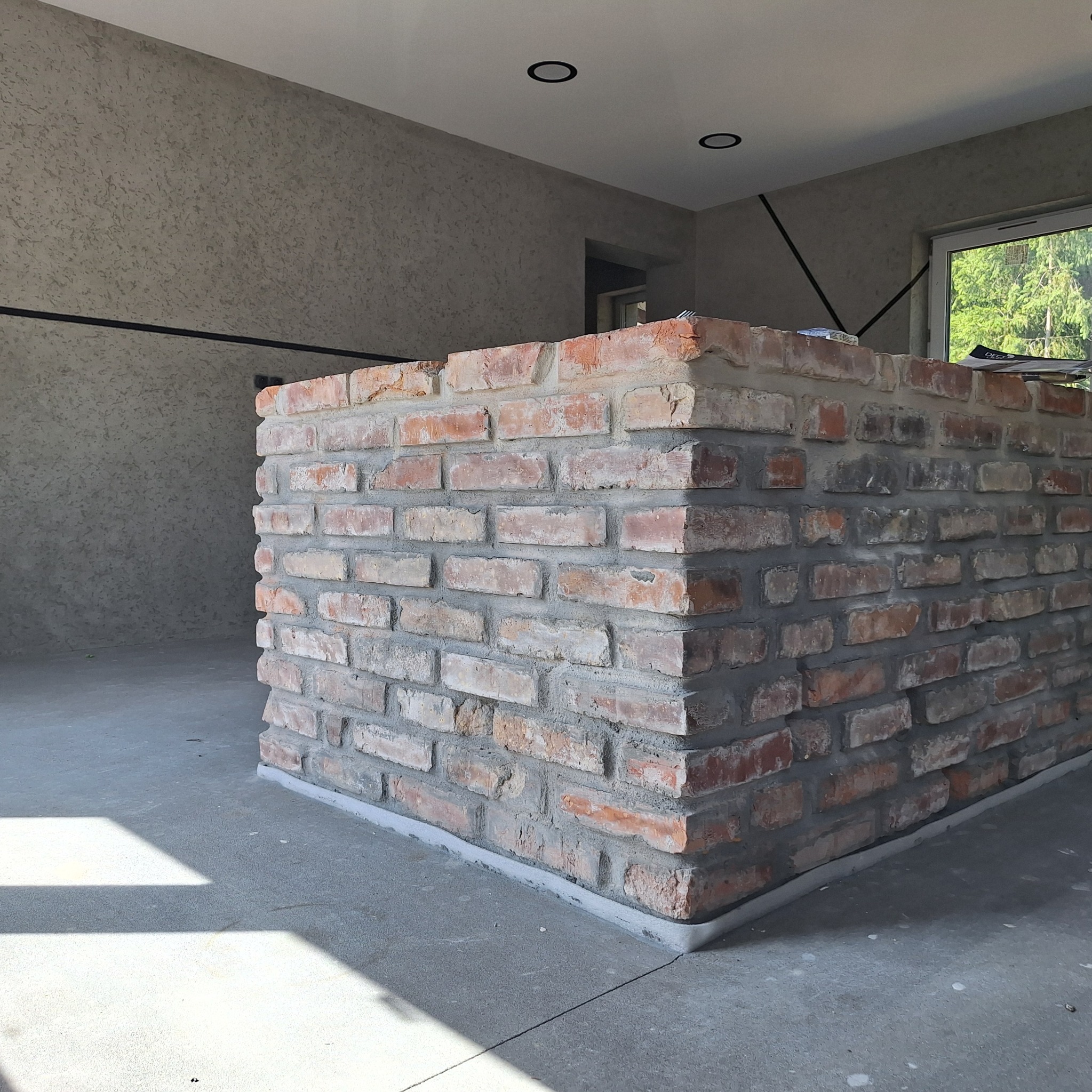 Murowana z cegły ściana działowa w surowym stanie, widok z dołu. Ściana z czerwonej cegły na tle szarej ściany z fakturą i okna. Widać fragment podłogi.