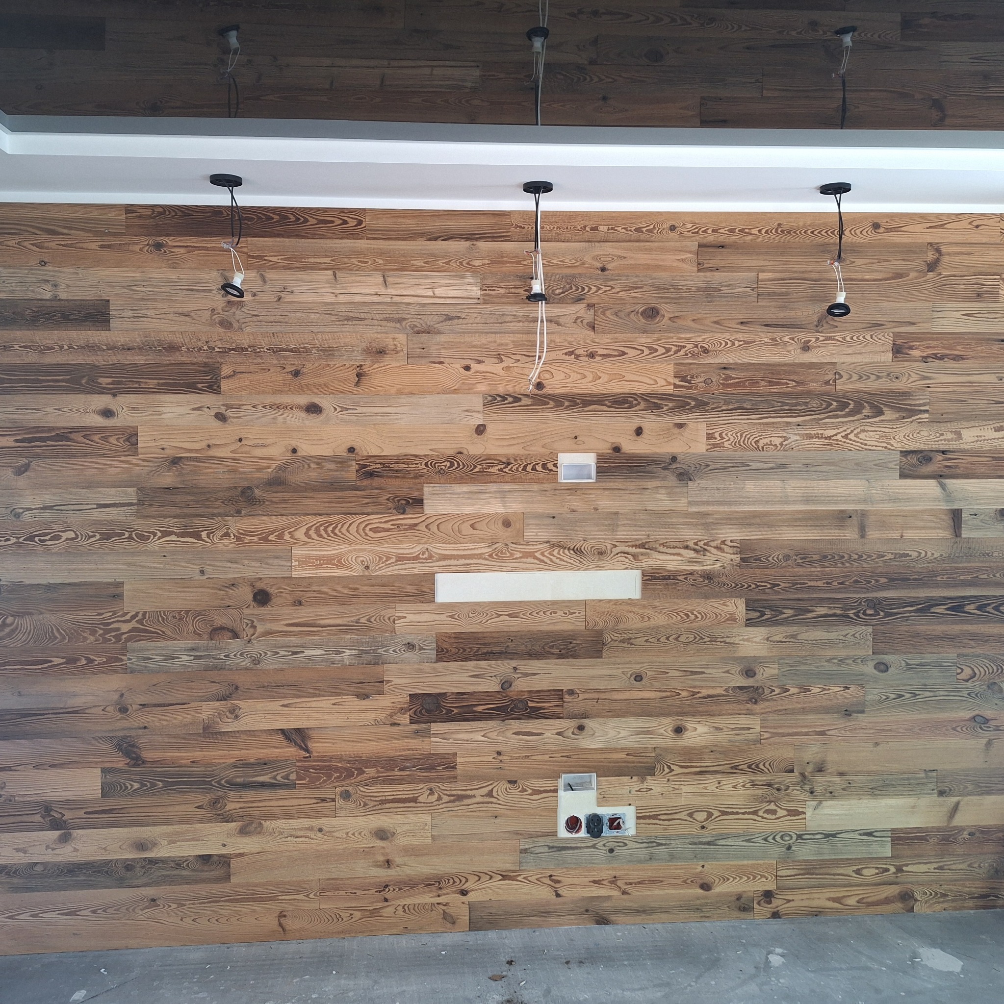 Ściana z drewnianych paneli o unikalnym usłojeniu, z widocznymi puszkami elektrycznymi i przewodami lamp, ukazująca etap prac wykończeniowych.