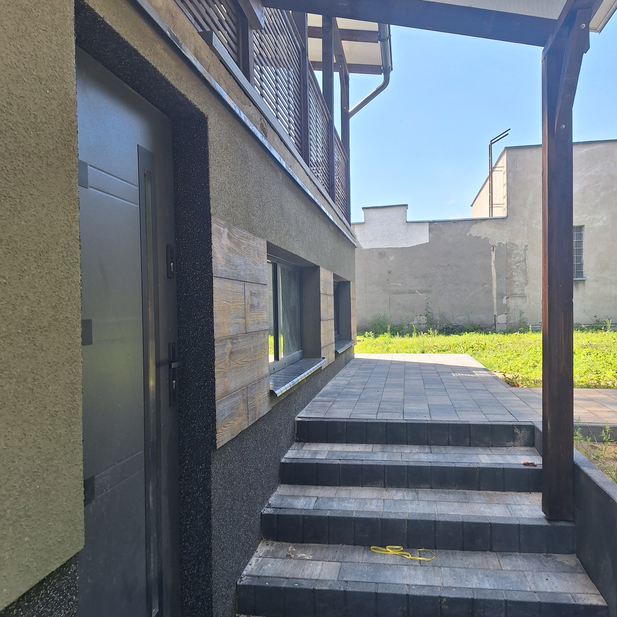 Elewacja domu z oknami obłożonymi panelami imitującymi drewno, schody wejściowe z kostki brukowej, fragment balkonu z drewnianą balustradą i widok na niebo.