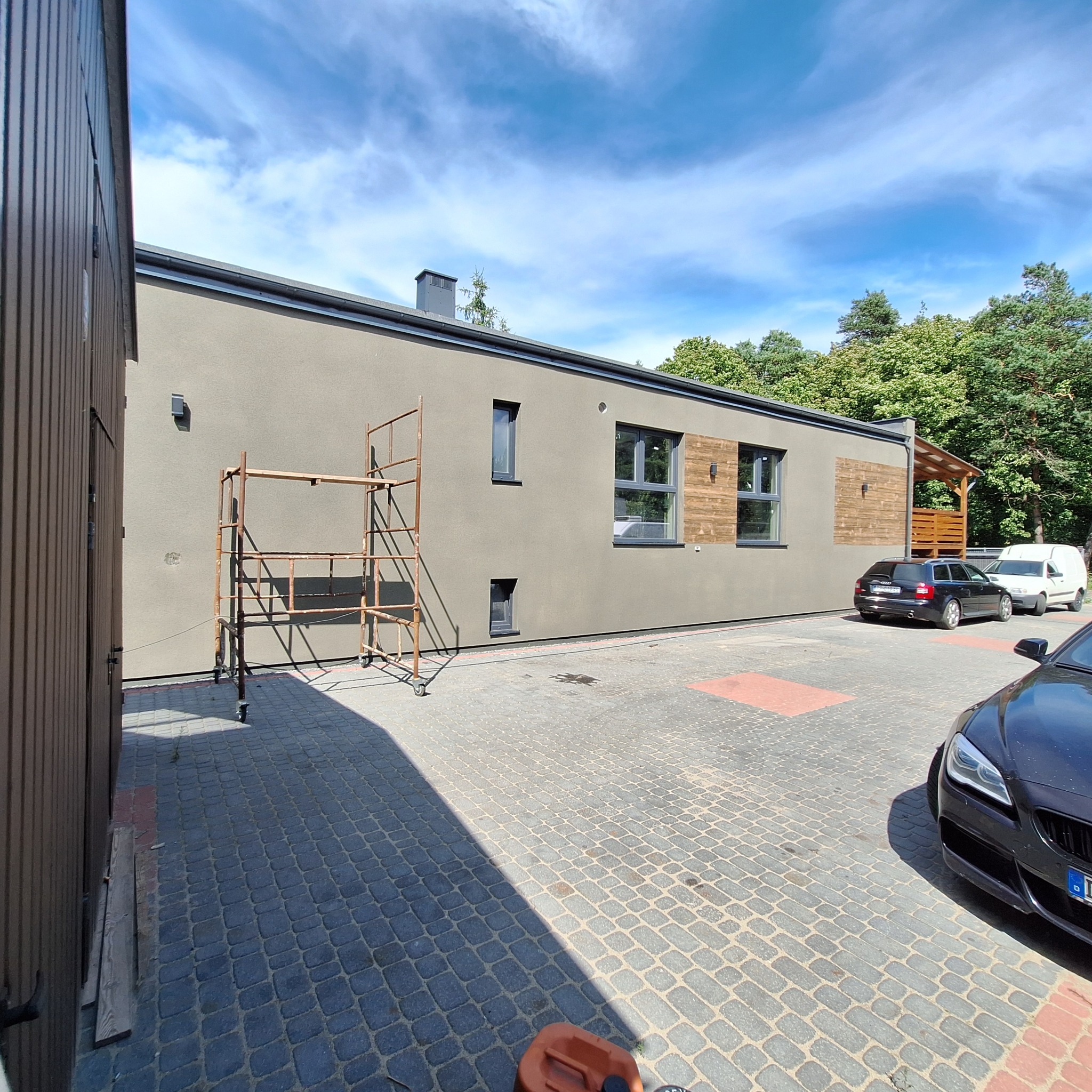 Elewacja budynku po szpachlowaniu i malowaniu na szaro, z drewnianymi akcentami. Widoczne rusztowanie, samochody i kostka brukowa. Słoneczny dzień.