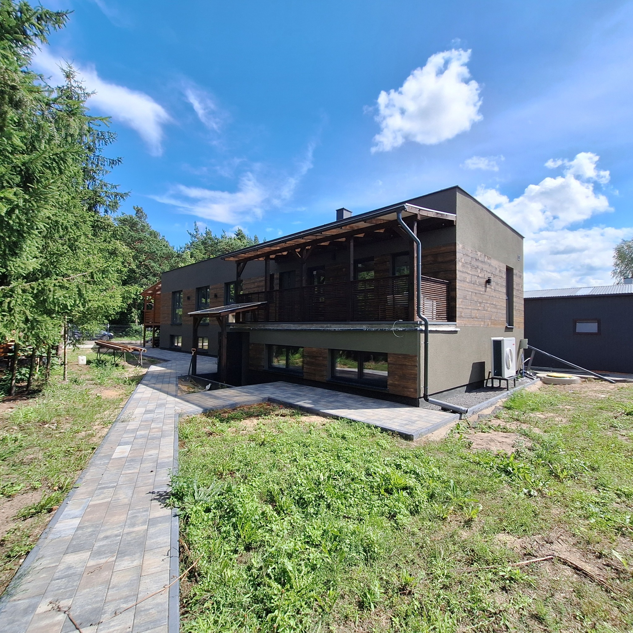 Dwukondygnacyjny dom z drewnianą elewacją i zadaszonym balkonem, otoczony zielenią i brukowanym chodnikiem. Widok z zewnątrz w słoneczny dzień.