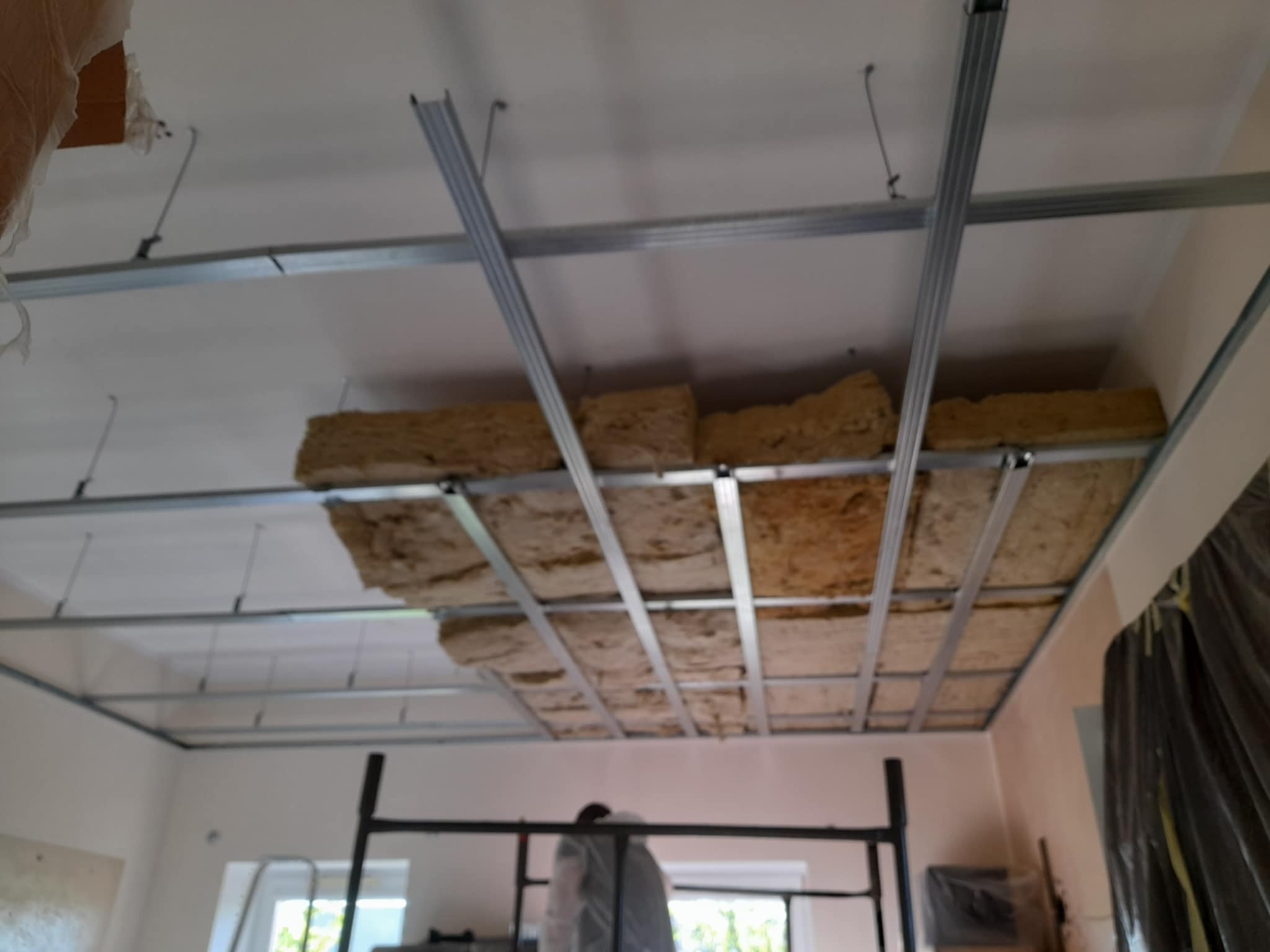 Montaż izolacji z wełny mineralnej w metalowym stelażu sufitu podwieszanego. Widoczne profile i fragment rusztowania.
