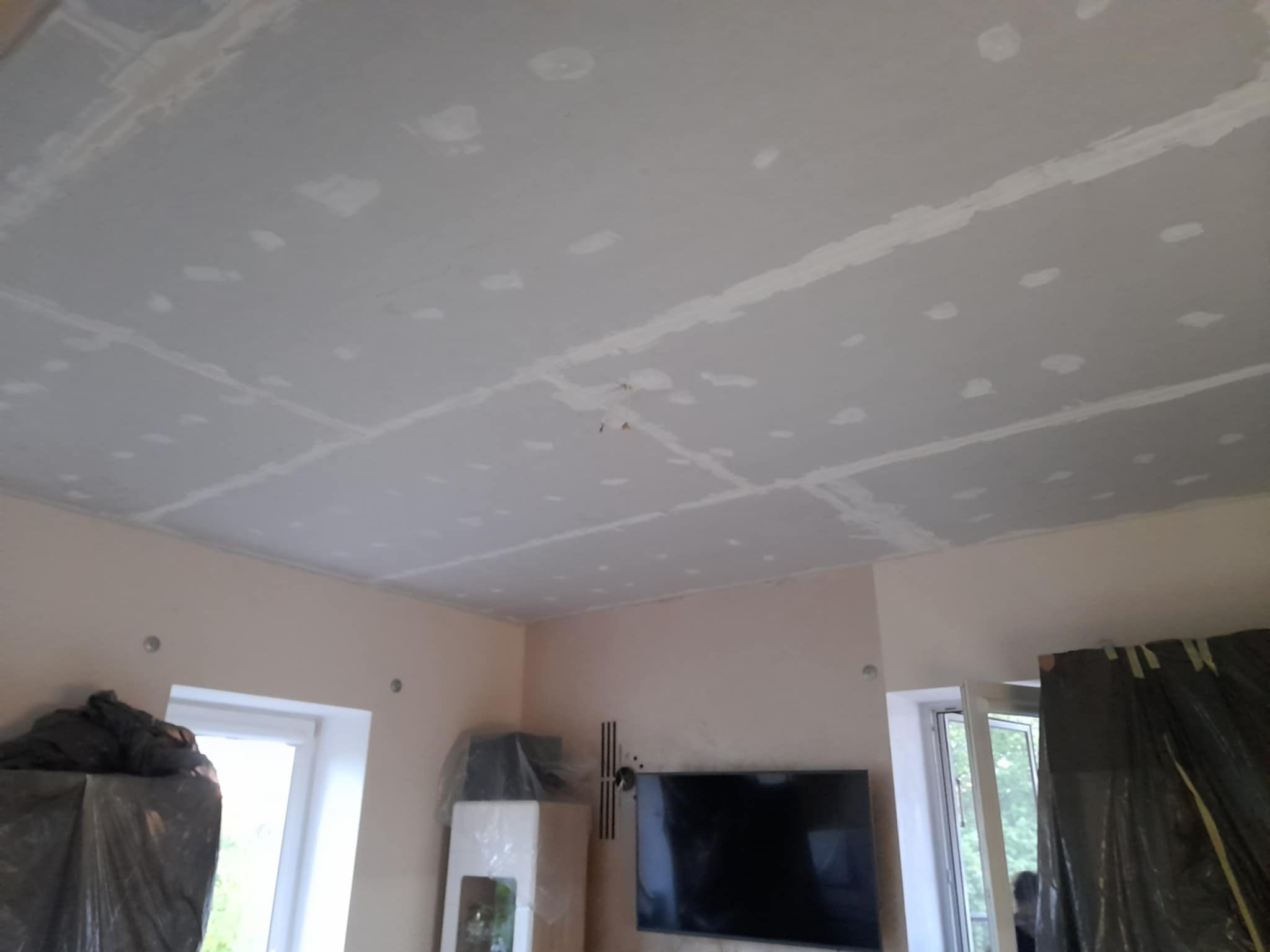 Sufit z płyt karton-gips, szpachlowany, z widocznymi łączeniami i śladami po szpachlowaniu. W tle fragment pokoju z oknem i telewizorem.