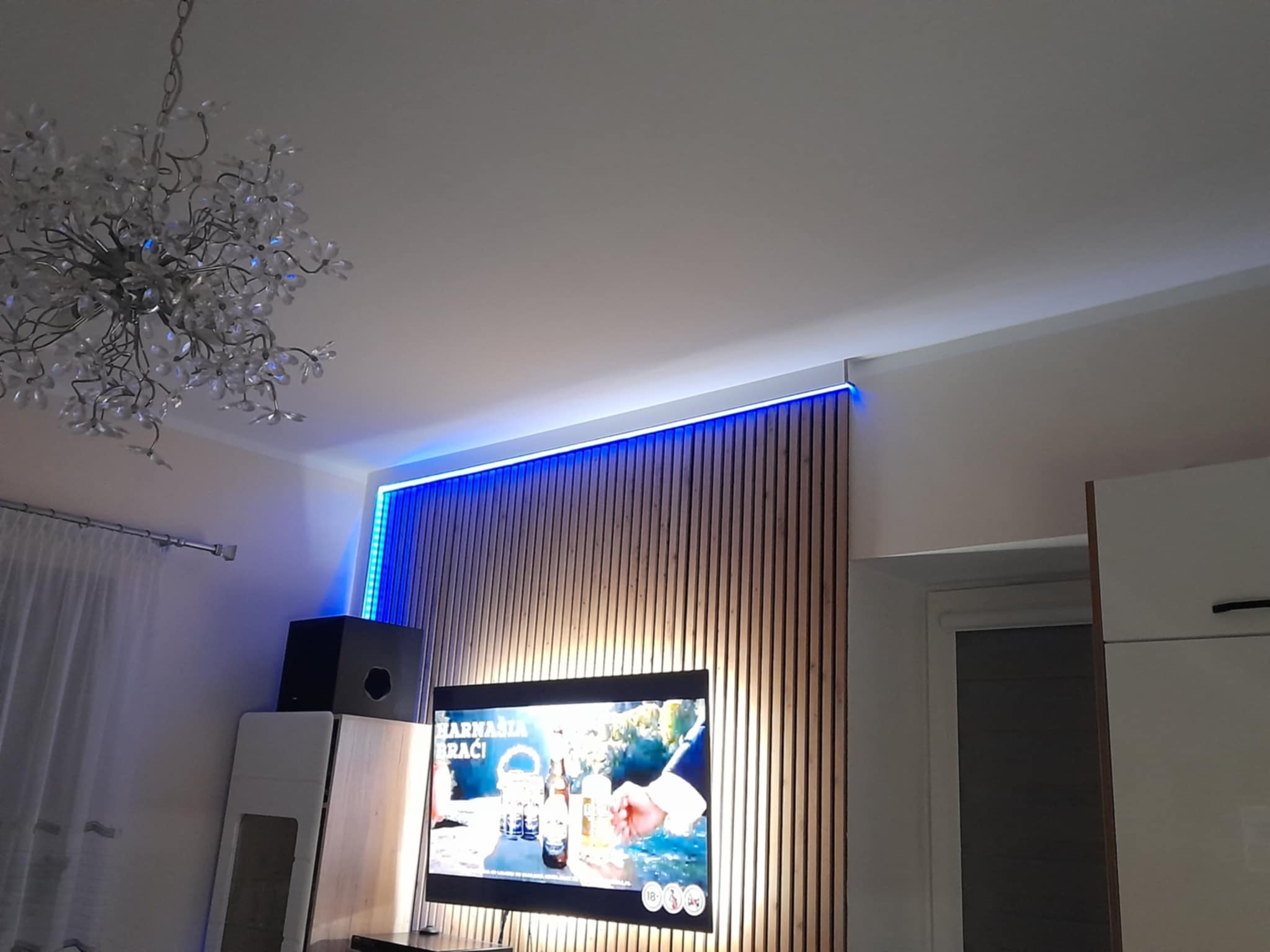 Nowoczesny salon z drewnianą ścianą lamelową podświetloną niebieskim LED, telewizor, kryształowy żyrandol i białe meble w minimalistycznym stylu.