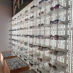 OPTYK KATOWICE Optyk.com OPTOMETRYSTA - Ekspozycja okularów na ścianie z ażurowym tłem i w gablotach, różnorodne modele w salonie optycznym. Plakat reklamowy z wizerunkiem osoby w tle.