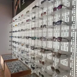 Ekspozycja okularów na ścianie z ażurowym tłem i w gablotach, różnorodne modele w salonie optycznym. Plakat reklamowy z wizerunkiem osoby w tle.