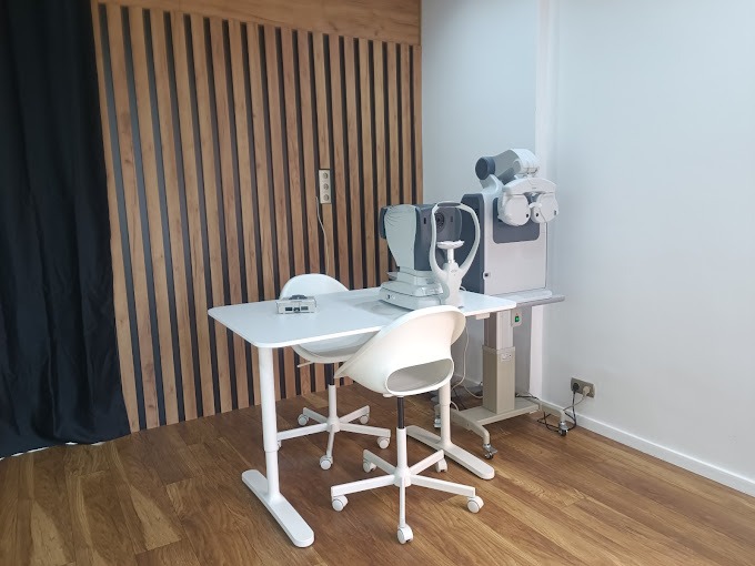 Nowoczesny gabinet okulistyczny z aparaturą do badania wzroku, białym biurkiem i krzesłami na tle drewnianej ściany i zasłony.