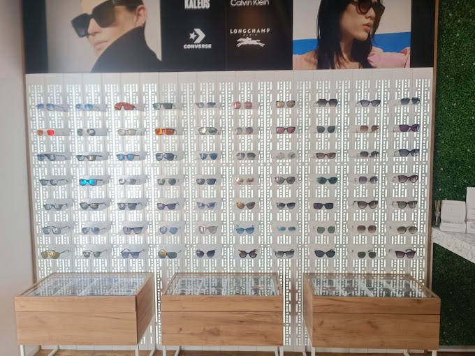 Ekspozycja okularów przeciwsłonecznych różnych marek na ażurowym tle, z drewnianymi gablotami na dole, w salonie optycznym z plakatami reklamowymi u góry.