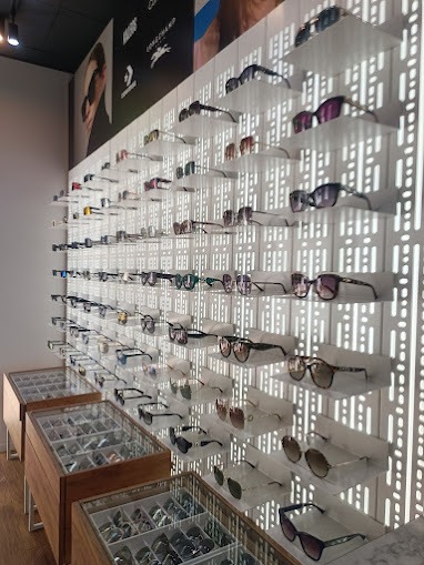 Ekspozycja okularów na ścianie z ażurowym tłem i w gablotach, różnorodne modele w salonie optycznym. Plakat reklamowy z wizerunkiem osoby w tle.