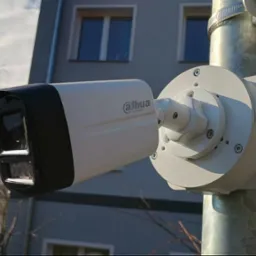 Kamera monitoringu Dahua na metalowym słupie, z budynkiem w tle. Widoczne detale montażu i obudowy kamery. Ujęcie z dołu, podkreślające wysokość instalacji.