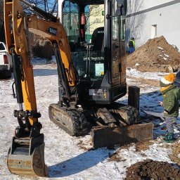 J&A SPÓŁKA Z OGRANICZONĄ ODPOWIEDZIALNOŚCIĄ - Kompaktowa koparka SANY z łyżką na śniegu, obok dziecko w zimowej kurtce i czapce z pomponem. W tle budynek i pracownik w odblaskowej kamizelce.