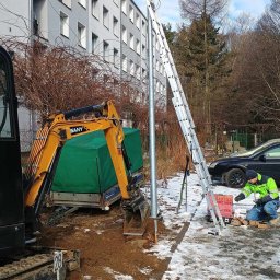 J&A SPÓŁKA Z OGRANICZONĄ ODPOWIEDZIALNOŚCIĄ - Montaż słupa oświetleniowego: koparka SANY, drabina oparta o słup, pracownik w odblaskowej kurtce przygotowuje elementy montażowe. Prace ziemne w toku.