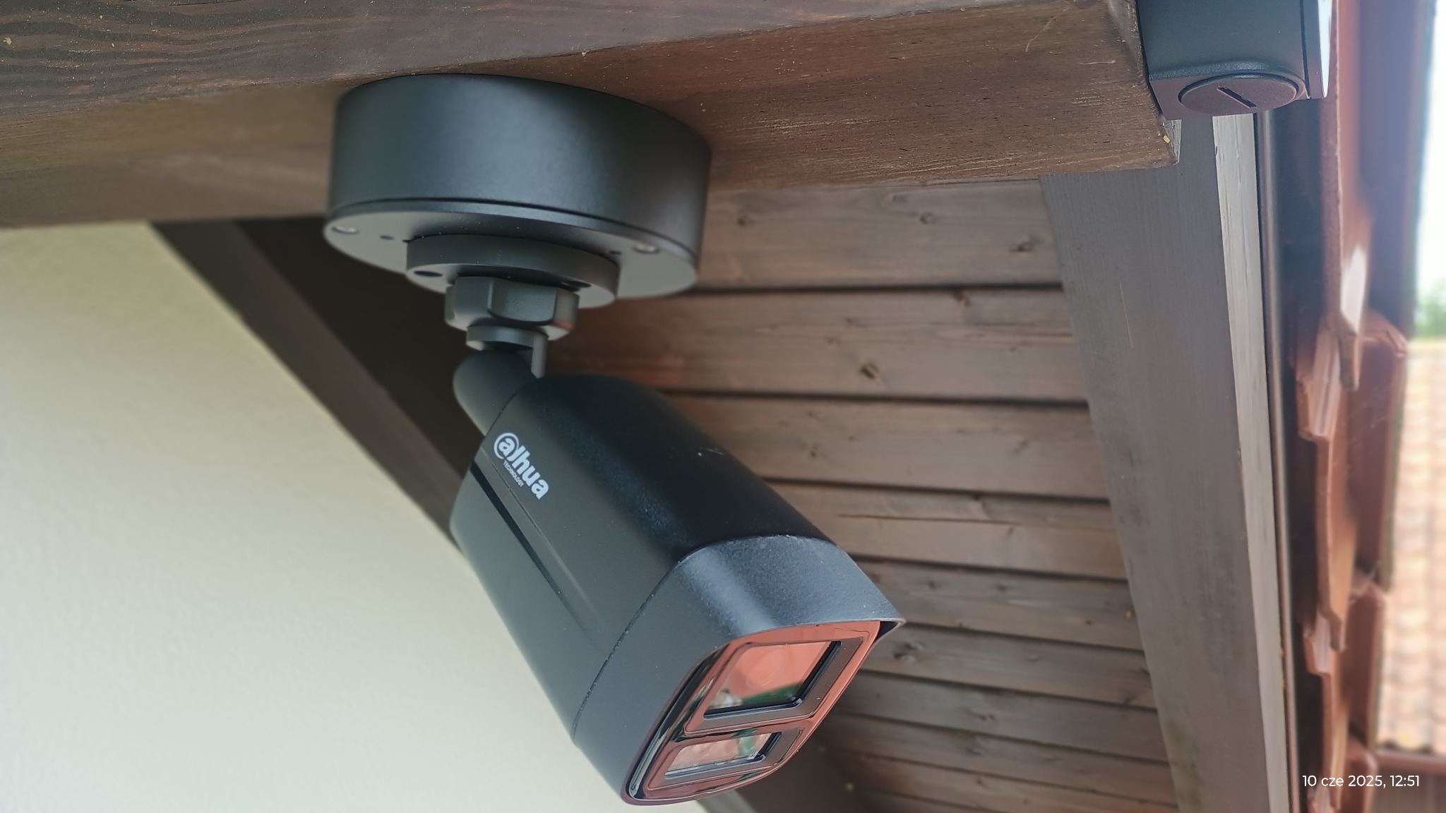 Czarna kamera Dahua zamontowana pod drewnianym okapem, widok z dołu, z widocznym logo i elementami mocującymi.
