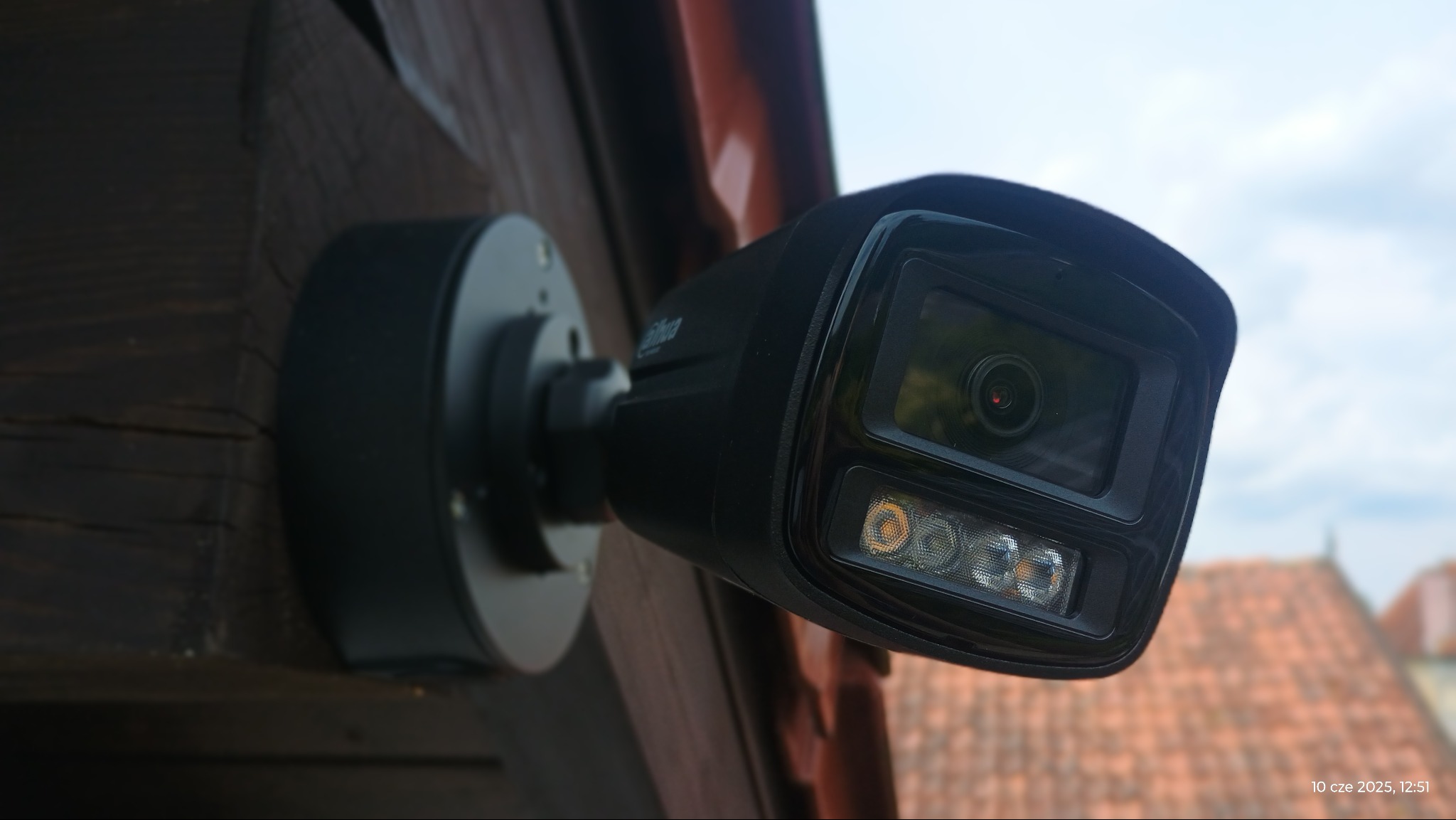 Czarna kamera monitoringu Dahua, zamontowana na drewnianej elewacji. Widoczne diody LED, obiektyw i fragment dachu w tle. Ujęcie z dołu, na zewnątrz budynku.