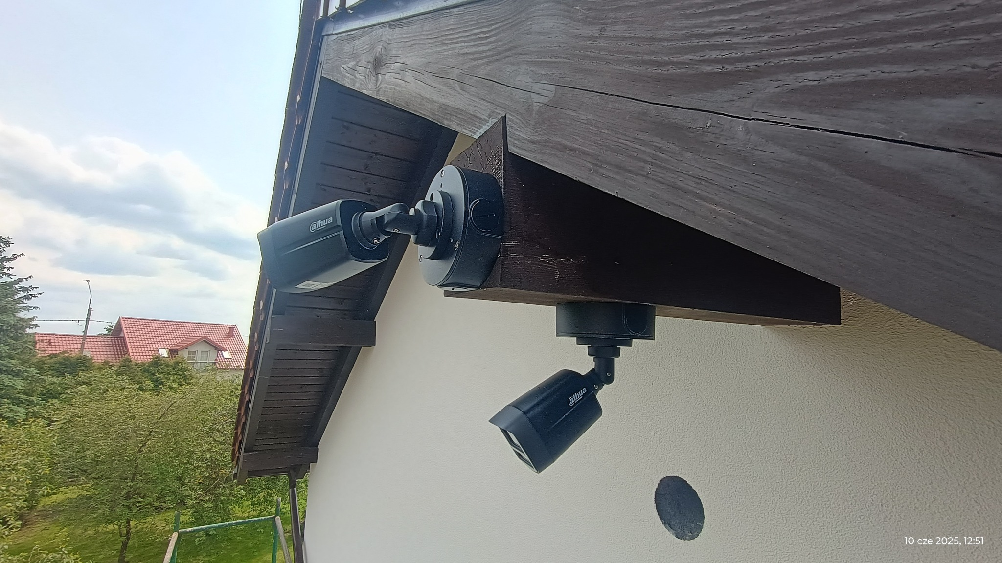 Dwie kamery monitoringu Dahua zamontowane na drewnianej podbitce dachu, skierowane w różnych kierunkach. Widok z dołu, na tle jasnej elewacji i fragmentu ogrodu.
