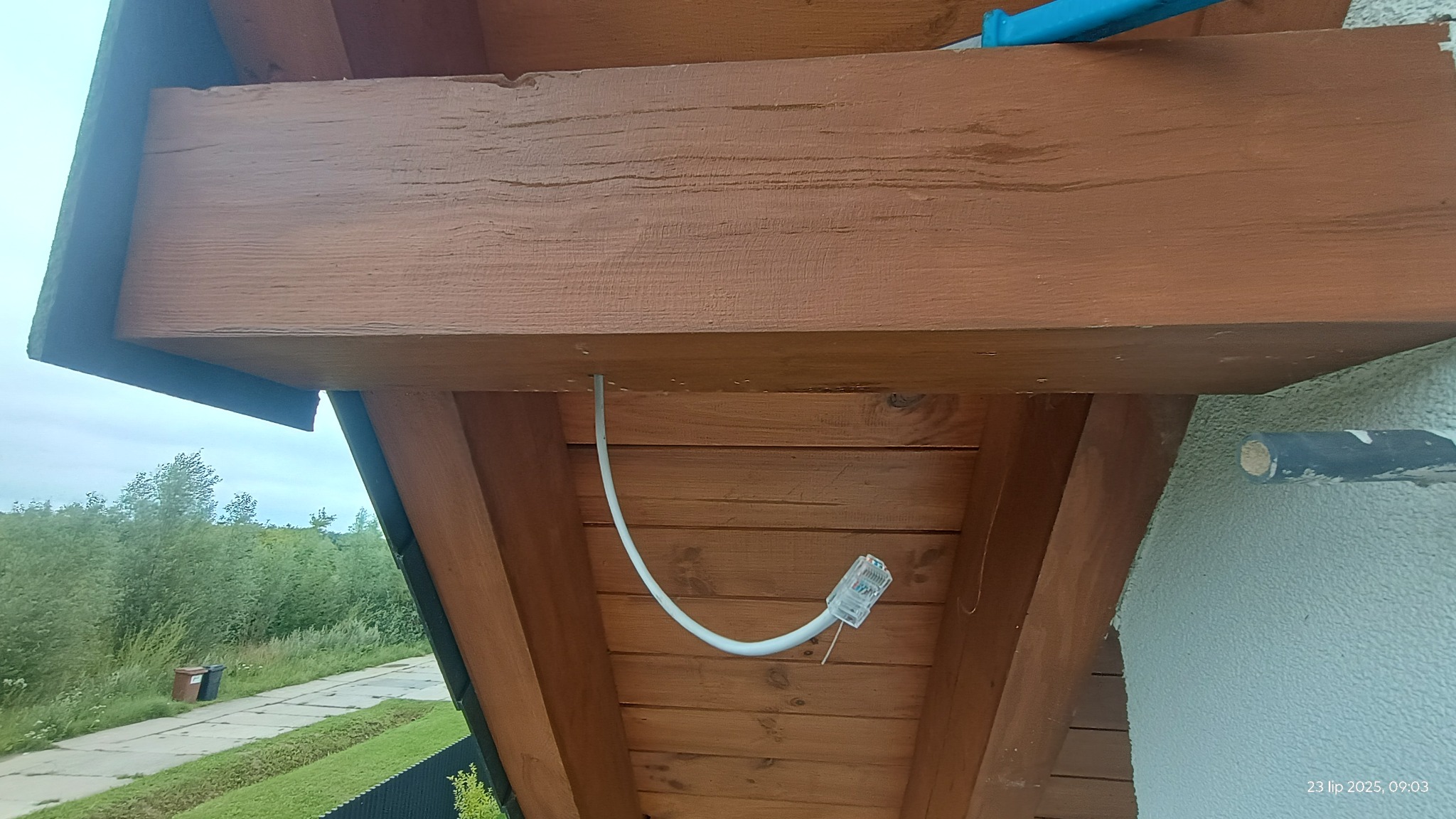 Biały kabel sieciowy z wtyczką RJ45 przymocowany do drewnianej konstrukcji dachu na zewnątrz budynku, z widocznym fragmentem elewacji i zieleni w tle.