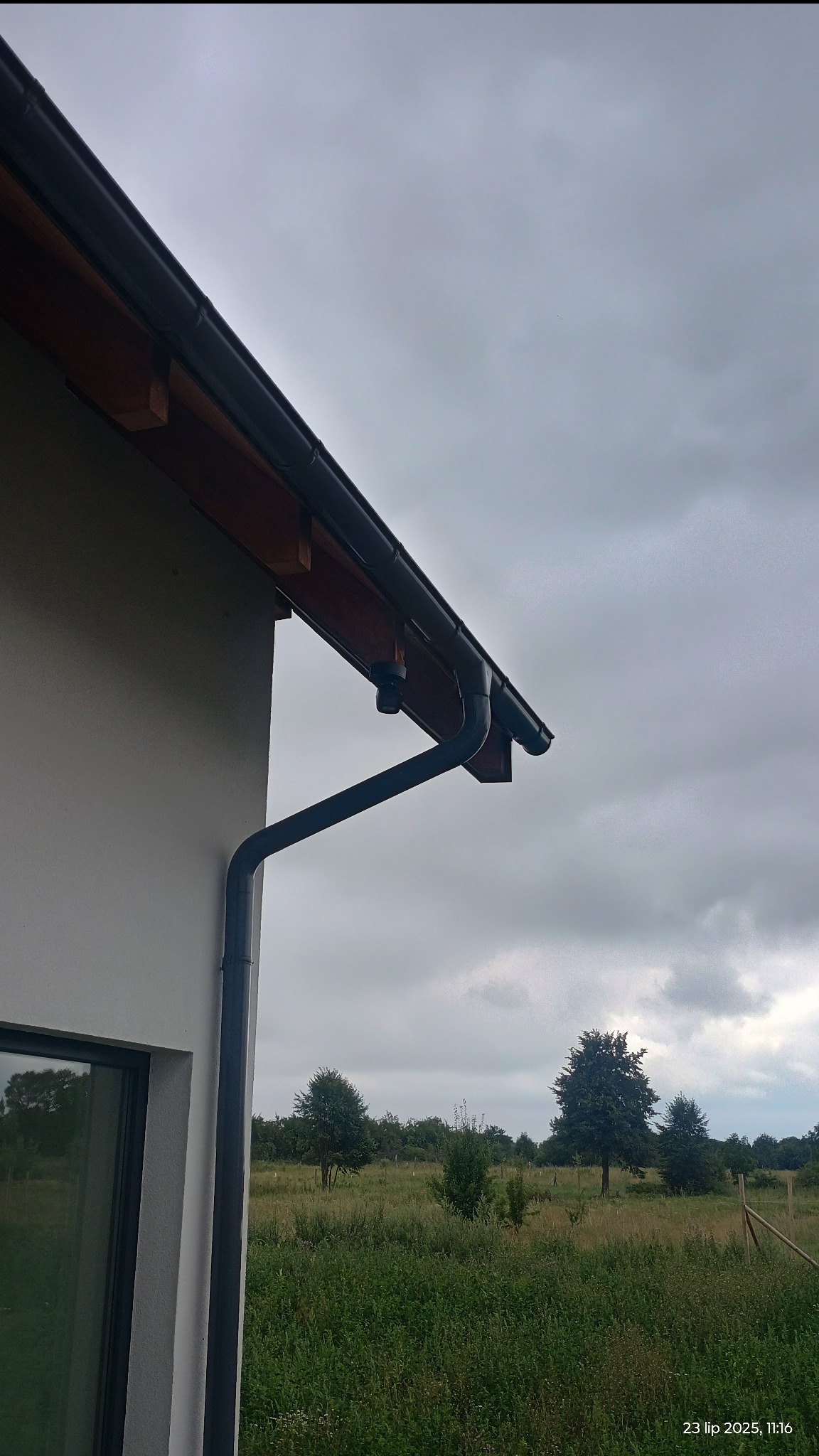 Kamera monitoringu zamontowana pod okapem domu z czarnymi rynnami, na tle pochmurnego nieba i łąki z drzewami. Widok z dołu.