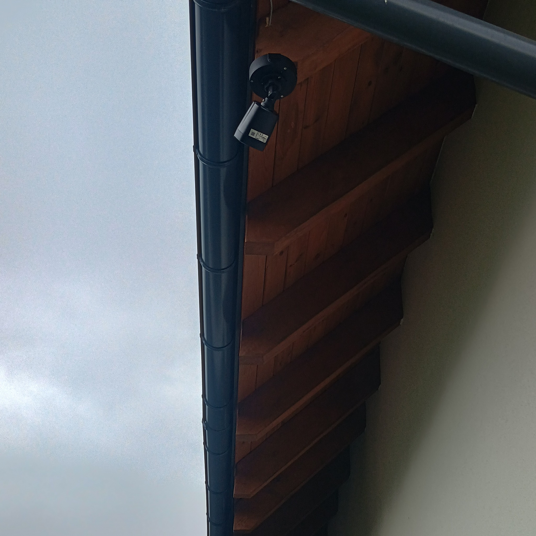 Czarna kamera monitoringu zamontowana pod drewnianym okapem, obok rynny. Widok z dołu na szare niebo.