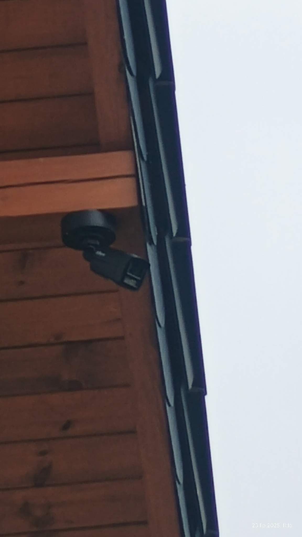Kamera monitoringu przymocowana do drewnianej elewacji budynku, widoczna z dołu, obok rynny. Urządzenie w kolorze czarnym.