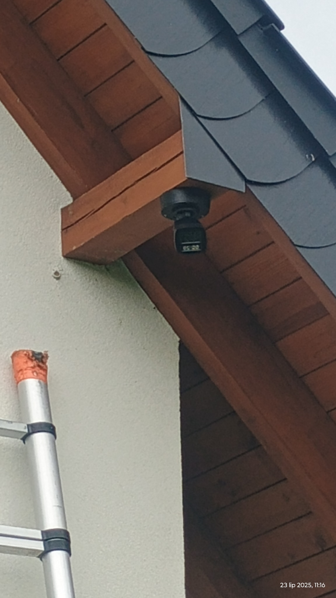 Czarna kamera monitoringu przymocowana do drewnianej podbitki dachowej, widoczny fragment drabiny oparty o jasną ścianę budynku.